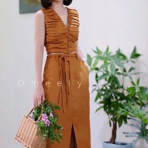 Peut inclure: Un ensemble deux pi&egrave;ces couleur tabac compos&eacute; d'un haut sans manches &agrave; col en V fronc&eacute; avec boutons et d'une jupe longue taille haute assortie avec ceinture tress&eacute;e, fente avant et poches lat&eacute;rales. Le texte Omeely est visible.