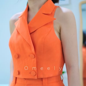 May include: A sleeveless orange cropped blazer with a double breasted closure and two buttons. The blazer is paired with matching high waisted orange pants. The text 'O meely' is visible on the front of the blazer.