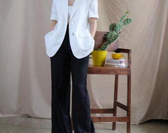 Pode incluir: Um blazer branco de manga curta com dois botões frontais e bolsos com aba, combinado com um top curto branco e calças pretas de perna larga. O visual é complementado por óculos de sol pretos e sapatos brancos, mantendo um estilo neutro.