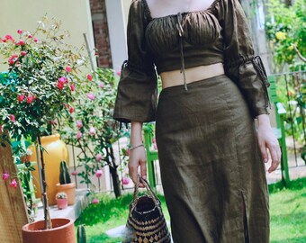 May include: An olive green two-piece set consisting of a cropped, off-the-shoulder top with gathered puff sleeves and a matching high-waisted midi skirt featuring a side slit. The set is paired with a woven basket bag and white ankle boots.