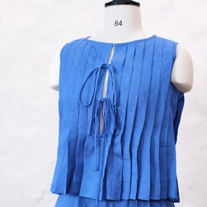 May include: A sleeveless blue top featuring vertical pleats and a front opening secured with two sets of thin fabric ties. The garment is displayed on a mannequin marked with the number 84, and the word Omeely is printed on the lower section.