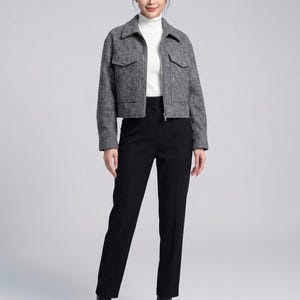 May include: A grey textured jacket with a collar, front zip, and two chest pockets, worn over a white mock neck top. The outfit includes black trousers and black ankle boots. The jacket features long sleeves and a cropped length.