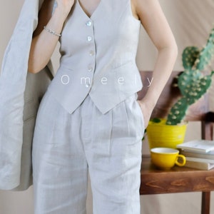 Puede incluir: Un chaleco beige con escote en V, cinco botones claros y dos peque&ntilde;os bolsillos ribeteados, combinado con pantalones de cintura alta a juego con pliegues y bolsillos laterales. El texto Omeely es visible en el centro del chaleco.