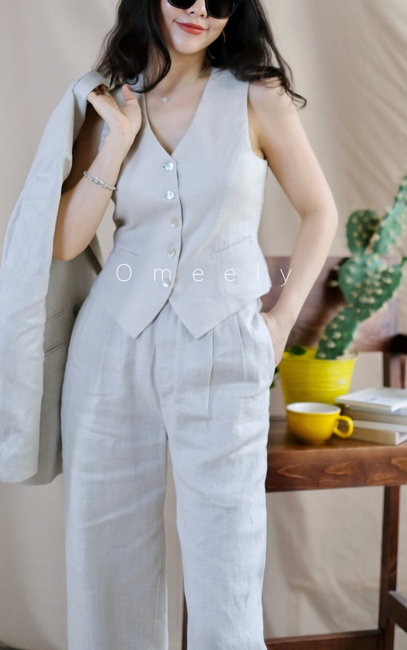 Puede incluir: Un chaleco beige con escote en V, cinco botones claros y dos peque&ntilde;os bolsillos ribeteados, combinado con pantalones de cintura alta a juego con pliegues y bolsillos laterales. El texto Omeely es visible en el centro del chaleco.