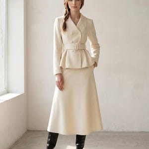 Op de afbeelding: Een crèmekleurige tweedelige set bestaande uit een double-breasted blazer met reverskraag, lange mouwen en een bijpassende riem met rechthoekige gesp, gecombineerd met een A-lijn midirok met hoge taille en zwarte leren kniehoge laarzen.