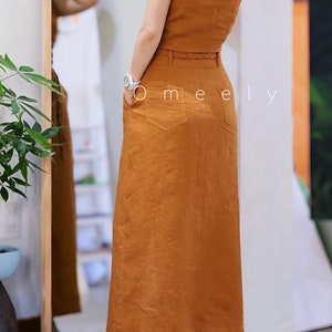 Peut inclure: Un haut sans manches couleur rouille et une jupe longue taille haute assortie avec une fine ceinture tress&eacute;e, deux poches arri&egrave;re et un ourlet droit. Le texte Omeely est visible sur la jupe. L'ensemble est port&eacute; avec des bottines rose clair.