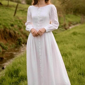 May include: A white, long-sleeved dress with a button-down front, worn with a straw hat. The dress is worn in a grassy field with a stream in the background. The dress has a fitted bodice and a flowing skirt. The dress is made of a light fabric.
