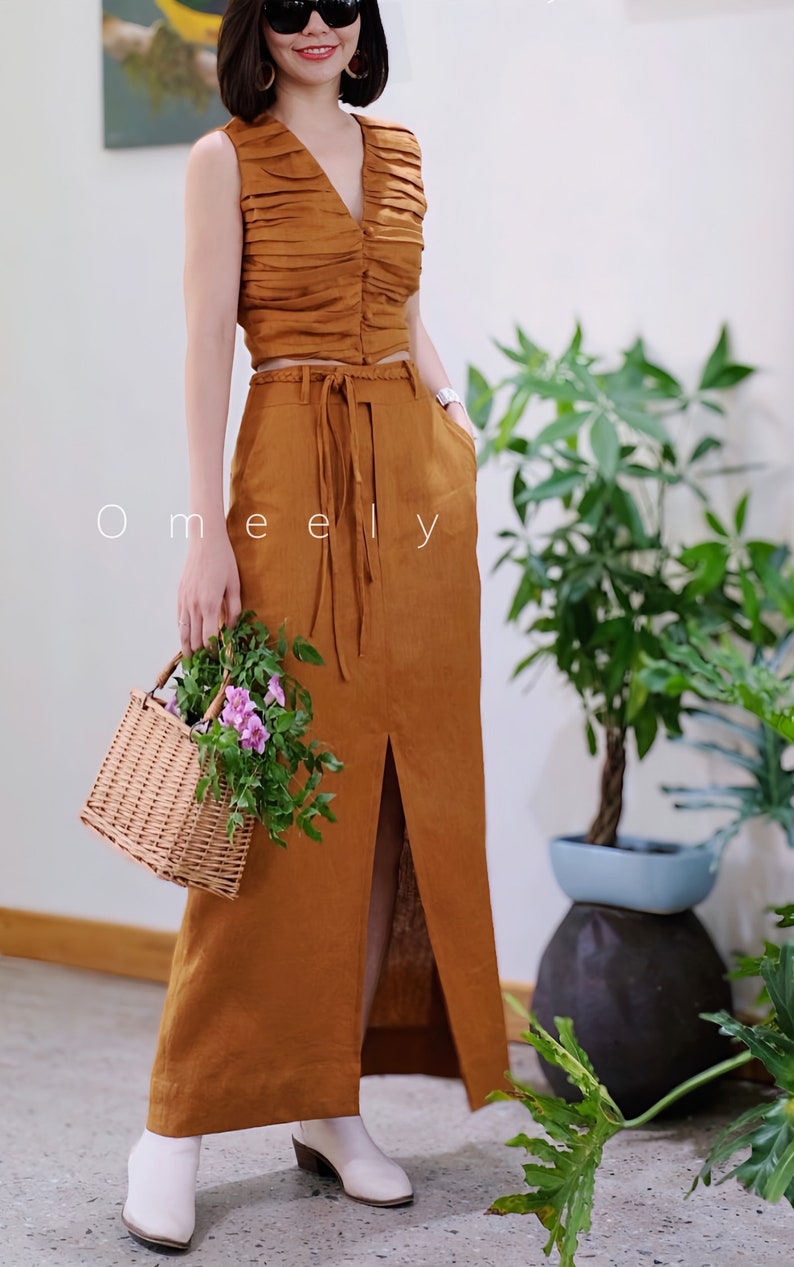 Peut inclure: Un ensemble deux pi&egrave;ces couleur tabac compos&eacute; d'un haut sans manches &agrave; col en V fronc&eacute; avec boutons et d'une jupe longue taille haute assortie avec ceinture tress&eacute;e, fente avant et poches lat&eacute;rales. Le texte Omeely est visible.