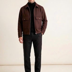 Può includere: Una giacca marrone con cerniera, colletto e due tasche frontali con patta, indossata sopra un maglione nero a collo alto. L'abbigliamento comprende pantaloni grigio antracite scuro e scarpe eleganti in pelle marrone, per un look coordinato.