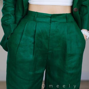 May include: A dark green linen trouser suit with a white top. The trousers have pleats and pockets. The jacket has a button fastening. The text "O meely" is visible on the trousers.