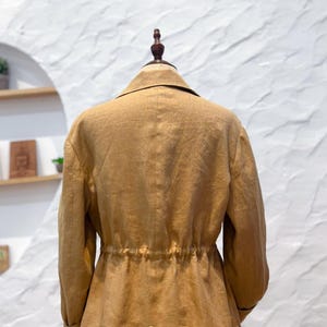 May include: A tan blazer with a collared neckline, long sleeves, and a cinched waist featuring a drawstring detail. The back of the garment displays a vertical seam and the text Omeely printed in white near the bottom hem.