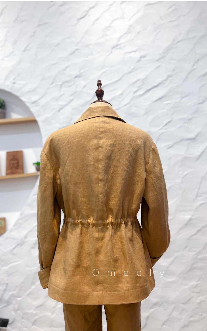 May include: A tan blazer with a collared neckline, long sleeves, and a cinched waist featuring a drawstring detail. The back of the garment displays a vertical seam and the text Omeely printed in white near the bottom hem.