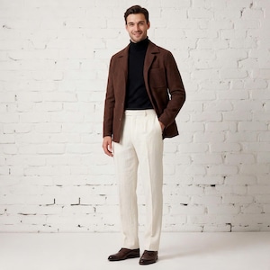 Può includere: Un blazer marrone con revers a dente e una tasca sul petto, indossato sopra un maglione a collo alto nero. L'outfit è completato da pantaloni bianco sporco con piega stirata e scarpe eleganti in pelle marrone scuro.