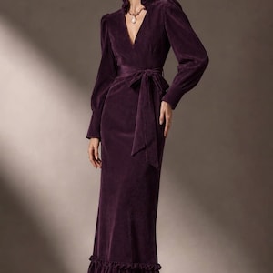 May include: A deep purple corduroy midi dress featuring a V-neckline, a ruffled collar, and long sleeves with cuffed wrists. The dress includes a matching fabric tie belt at the waist and a ruffled hemline, paired with dark brown pointed-toe heels.