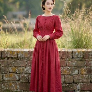 May include: A full-length, red dress with long sleeves and a button-down design. The dress features a round neckline and a flowing skirt. The person is standing in front of a brick wall and green grass.