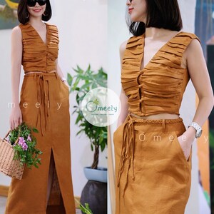 Peut inclure: Un ensemble deux pi&egrave;ces de couleur brun moutarde compos&eacute; d'un haut court sans manches &agrave; col en V avec des d&eacute;tails pliss&eacute;s horizontaux et des boutons sur le devant, assorti d'une jupe longue taille haute avec une fente et une ceinture tress&eacute;e.