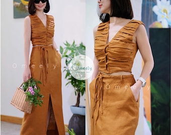 May include: A two-piece set in a mustard-brown color featuring a sleeveless, V-neck crop top with horizontal pleated detailing and front button closures, paired with a high-waisted maxi skirt that includes a front slit and a braided tie belt.