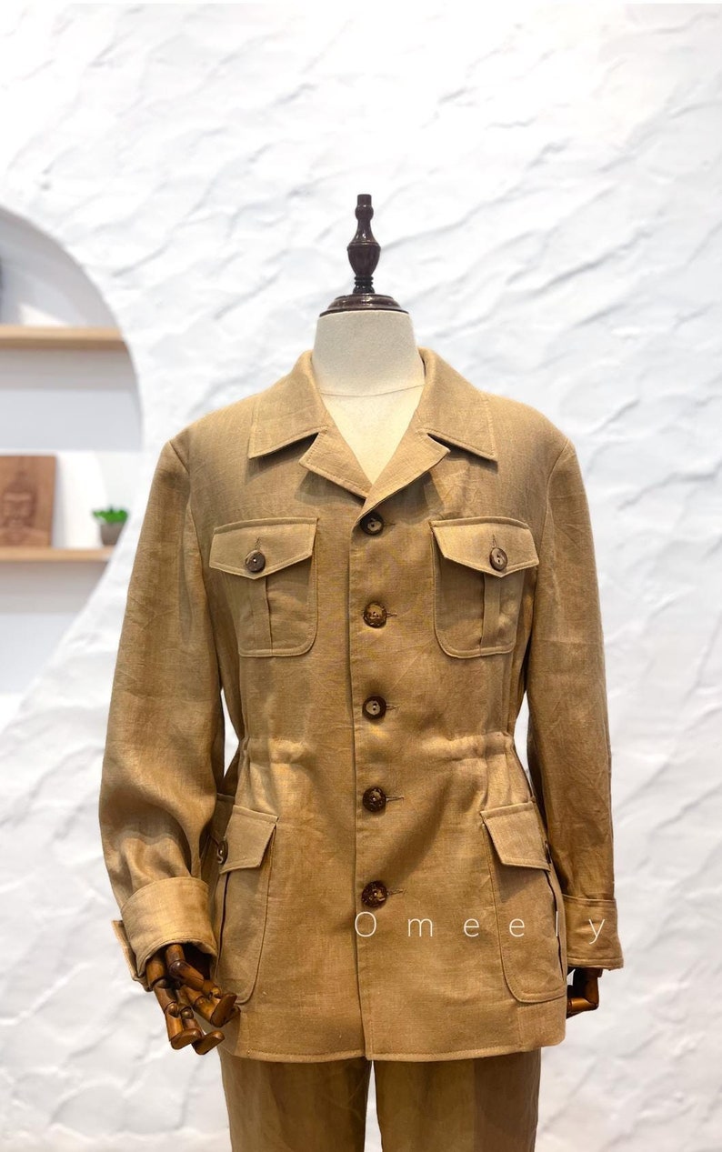 May include: A tan safari-style jacket featuring a collared neckline, long sleeves with buttoned cuffs, and a cinched waist. The jacket includes two buttoned chest pockets and two lower buttoned pockets, with a central button closure. The text Omeely is visible.