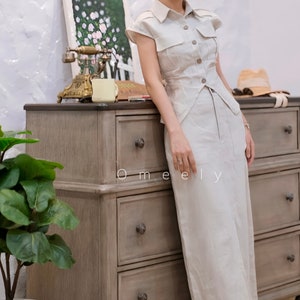 May include: A beige short-sleeved button-down shirt with a pointed collar and two chest pockets, paired with a matching long skirt. The outfit features a structured bodice with a buttoned front closure and a clean, straight-cut silhouette.