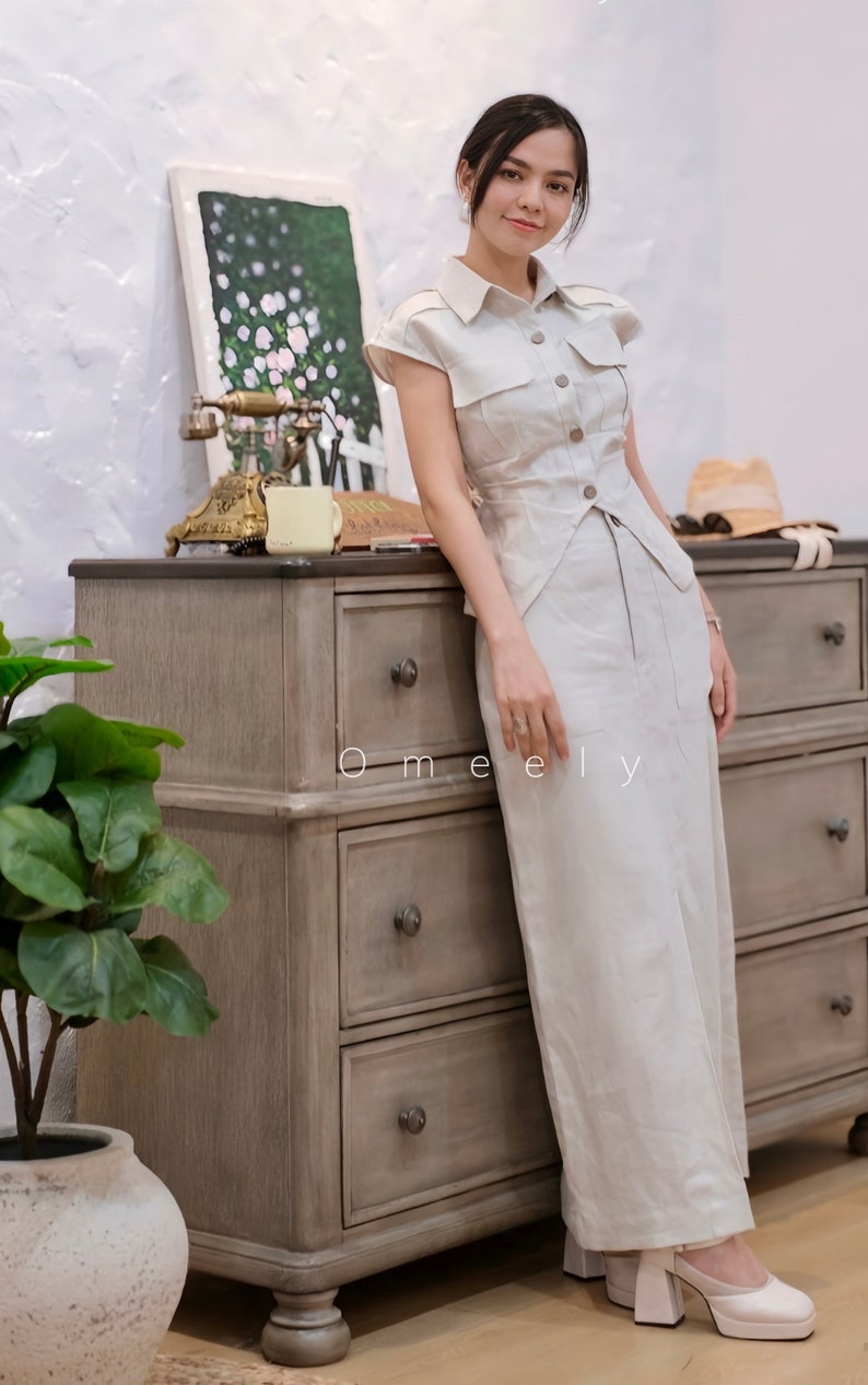 May include: A beige short-sleeved button-down shirt with a pointed collar and two chest pockets, paired with a matching long skirt. The outfit features a structured bodice with a buttoned front closure and a clean, straight-cut silhouette.
