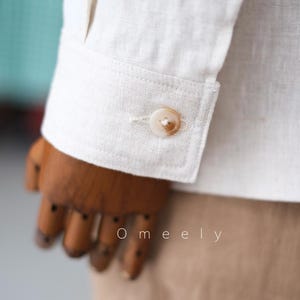 May include: A long-sleeved off-white shirt featuring a structured cuff with a single round, light brown marbled button. The fabric has a visible woven texture. The text Omeely is printed in white letters on the lower portion of the sleeve.