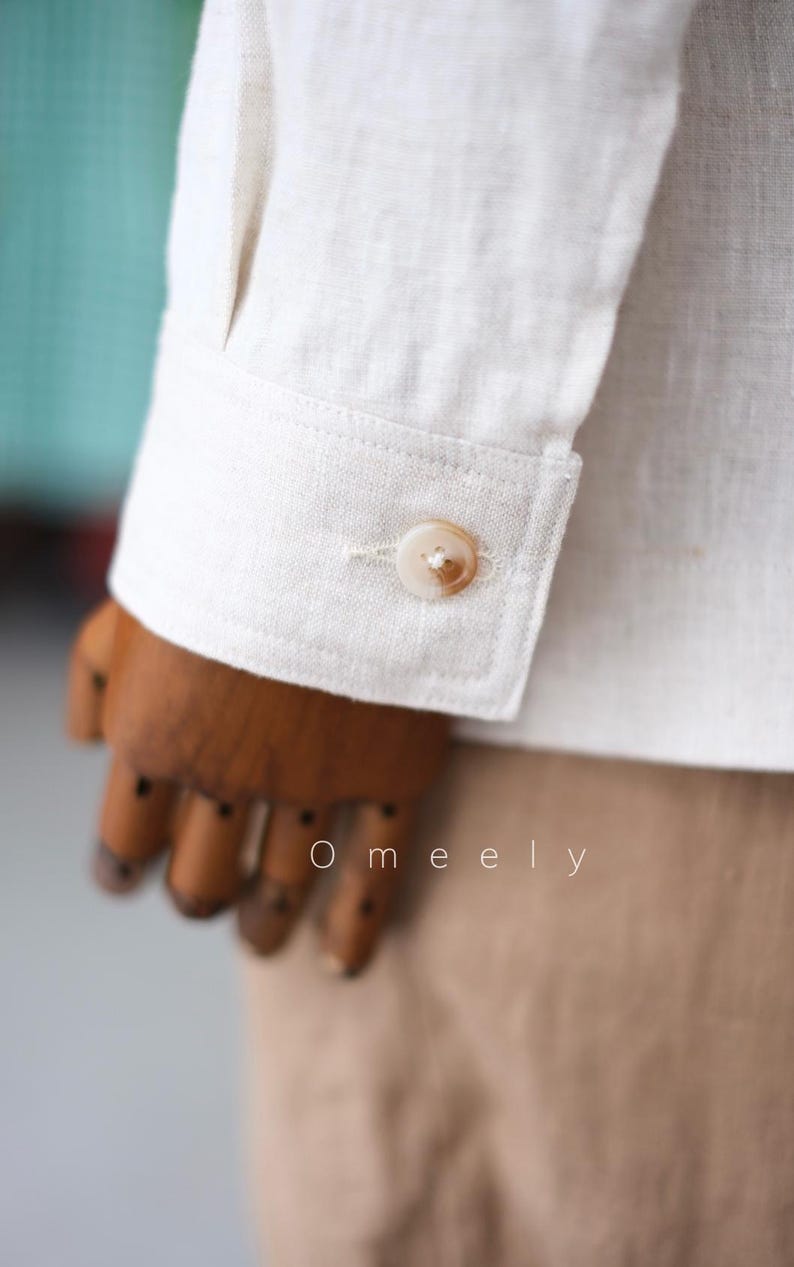May include: A long-sleeved off-white shirt featuring a structured cuff with a single round, light brown marbled button. The fabric has a visible woven texture. The text Omeely is printed in white letters on the lower portion of the sleeve.