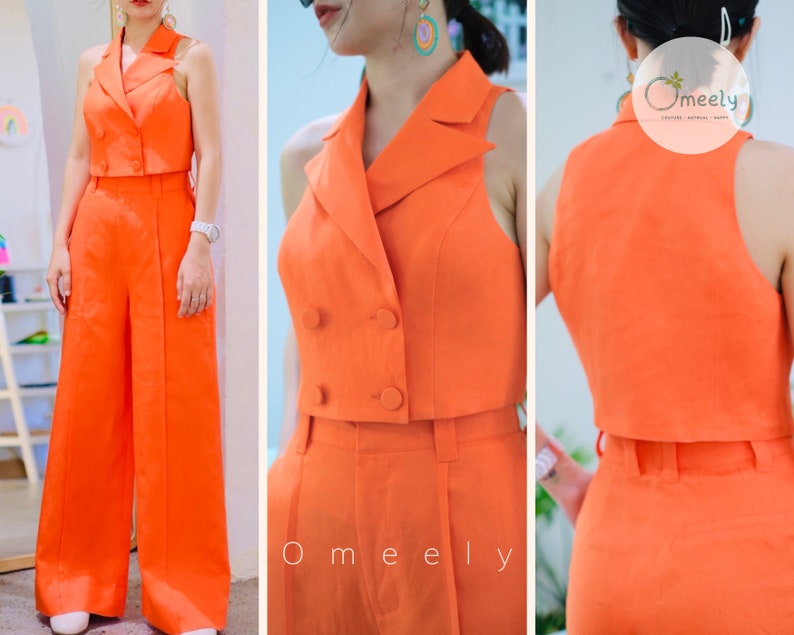 K&ouml;nnte beinhalten: Ein leuchtend orangefarbenes zweiteiliges Outfit mit einem kurzen Blazer-Oberteil und einer weiten Hose. Das Oberteil hat einen zweireihigen Verschluss und einen spitzen Kragen. Die Hose hat eine gerade Beinlinie und eine hohe Taille.