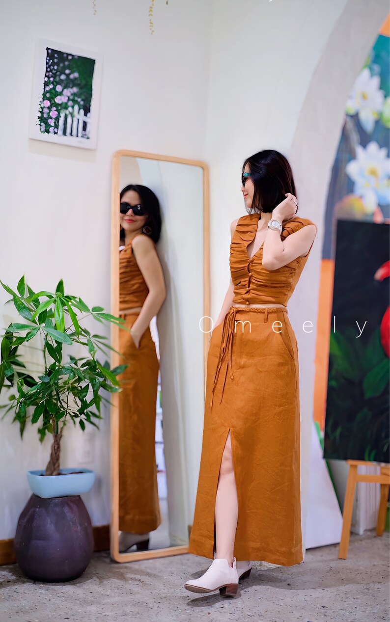 Peut inclure: Un ensemble deux pi&egrave;ces compos&eacute; d'un haut court sans manches, fronc&eacute; et boutonn&eacute;, et d'une jupe longue taille haute assortie de couleur rouille unie. La jupe pr&eacute;sente une fente sur le devant, des poches lat&eacute;rales et une fine ceinture tress&eacute;e.