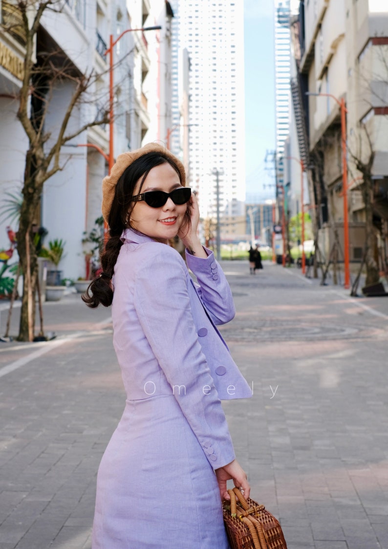 Pu&ograve; includere: Un completo composto da blazer e gonna color lavanda con chiusura a bottoni sulla giacca, abbinato a un basco color cuoio, occhiali da sole neri e una borsa a cestino intrecciata marrone. Il testo Omeely &egrave; stampato in bianco sul blazer.