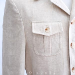 May include: A beige blazer with a textured finish, featuring a collared neckline, a single chest pocket with a buttoned flap, and a front button closure. The word Omeely is printed in white text on the lower left side of the garment.