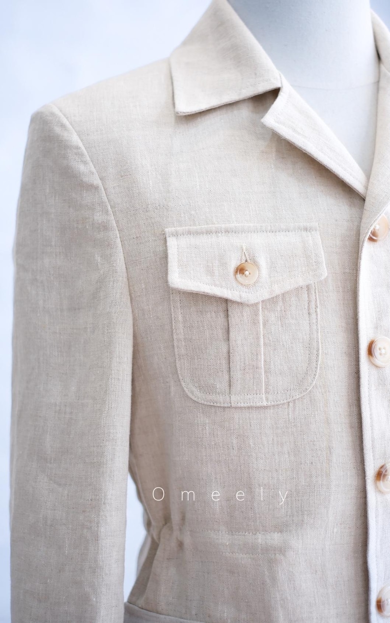 May include: A beige blazer with a textured finish, featuring a collared neckline, a single chest pocket with a buttoned flap, and a front button closure. The word Omeely is printed in white text on the lower left side of the garment.