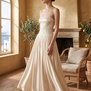 May include: A cream-colored, floor-length dress with spaghetti straps and a fitted bodice. The skirt flares out from the waist. The dress is worn with matching heels. The background features a window, a fireplace, and a wicker chair.