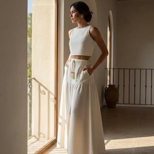 May include: A two-piece white outfit featuring a sleeveless crop top and a high-waisted maxi skirt with a front slit. The skirt has pockets and a drawstring waist. The outfit is made of a light-colored fabric and is suitable for warm weather.