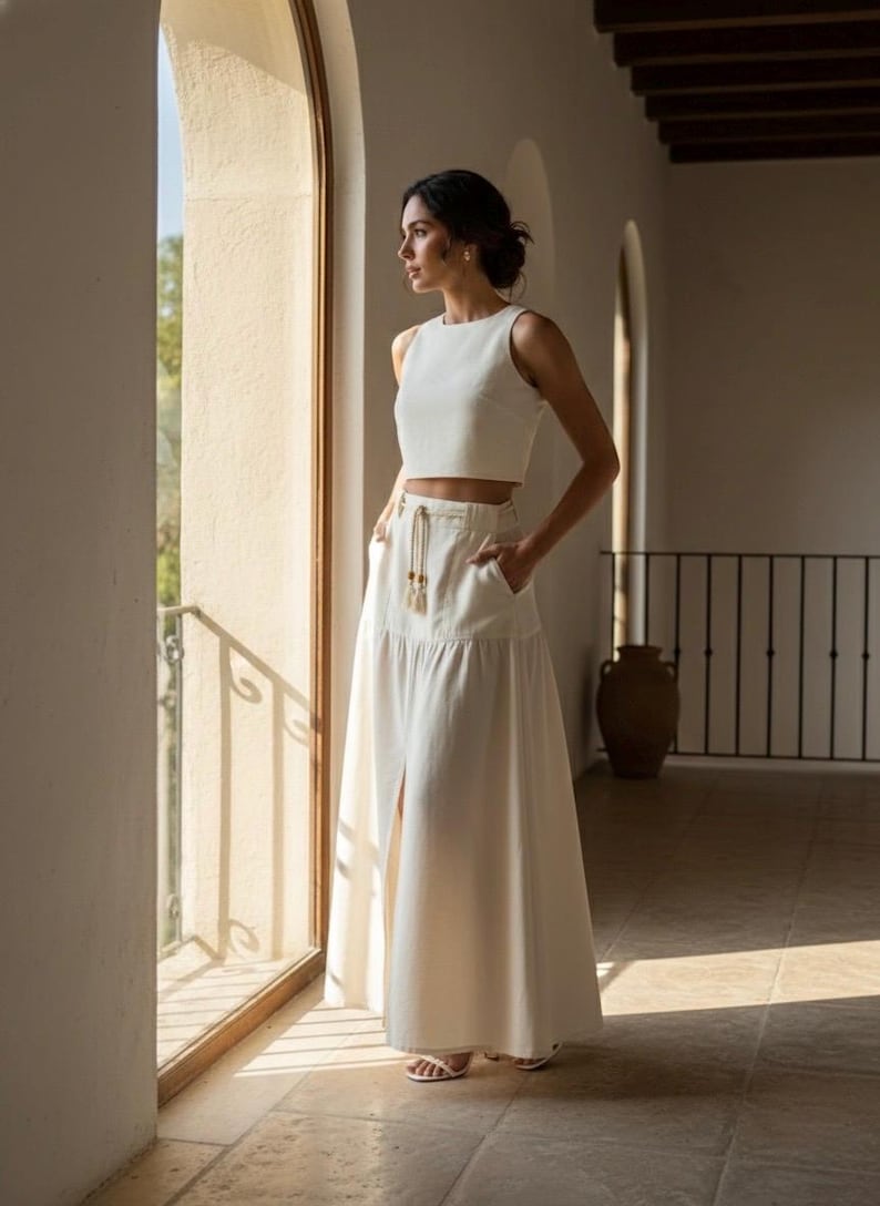 May include: A two-piece white outfit featuring a sleeveless crop top and a high-waisted maxi skirt with a front slit. The skirt has pockets and a drawstring waist. The outfit is made of a light-colored fabric and is suitable for warm weather.