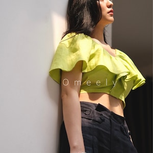 May include: A woman wearing a lime green off-the-shoulder crop top with ruffles and black high-waisted pants. She is holding a book in her hand.