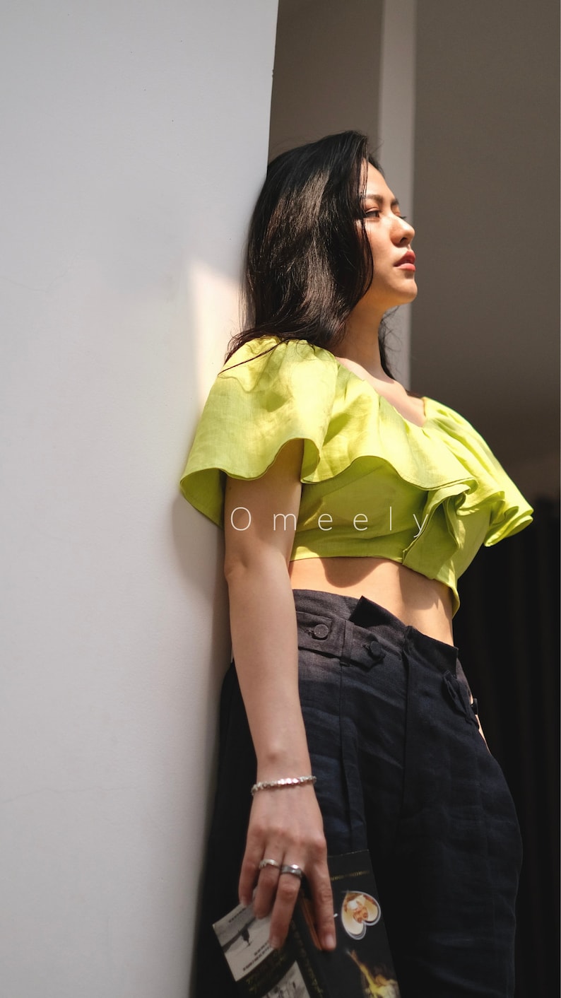 May include: A woman wearing a lime green off-the-shoulder crop top with ruffles and black high-waisted pants. She is holding a book in her hand.