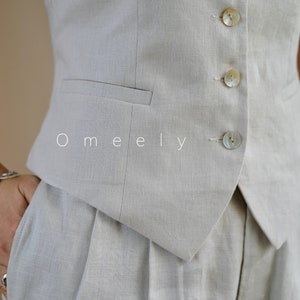 Puede incluir: Un chaleco sin mangas de color beige claro con dobladillo en punta, un bolsillo ribeteado y cuatro botones de n&aacute;car iridiscente en la parte delantera. La palabra Omeely est&aacute; impresa en blanco sobre la tela. El chaleco se combina con pantalones de pinzas a juego.