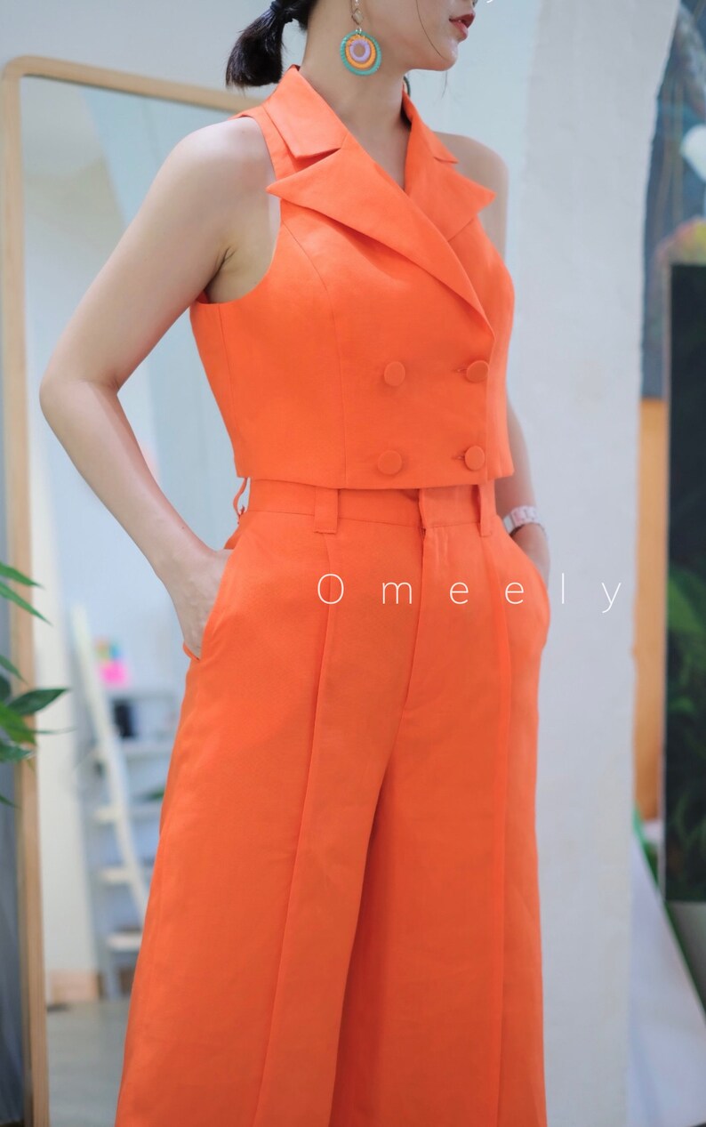May include: A sleeveless orange cropped blazer with a double breasted closure and a matching pair of wide leg pants. The pants have pleats in the front and pockets on the sides. The text 'O meely' is visible on the pants.