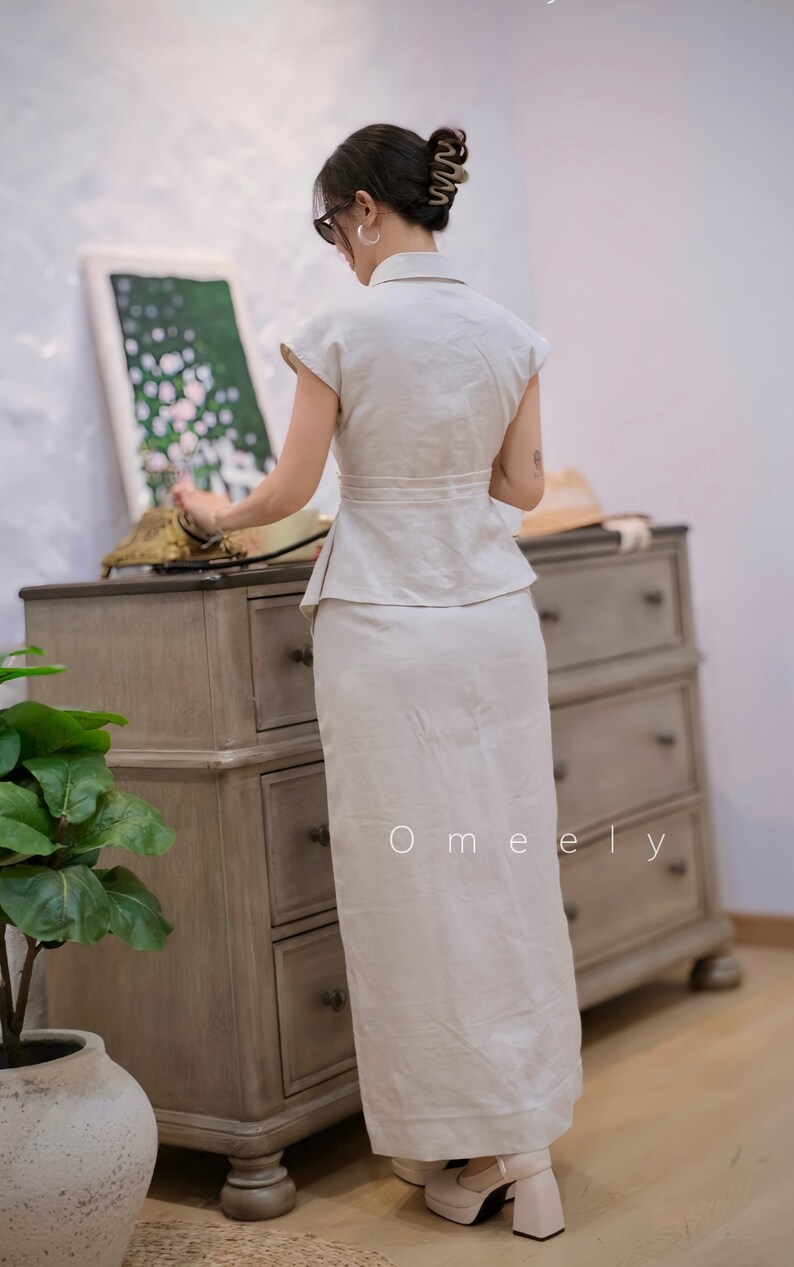 May include: A two-piece set consisting of a short-sleeved, collared peplum top and a matching floor-length skirt in a solid off-white color. The top features a cinched waist with a wide band, and the skirt has a straight, clean-lined silhouette.