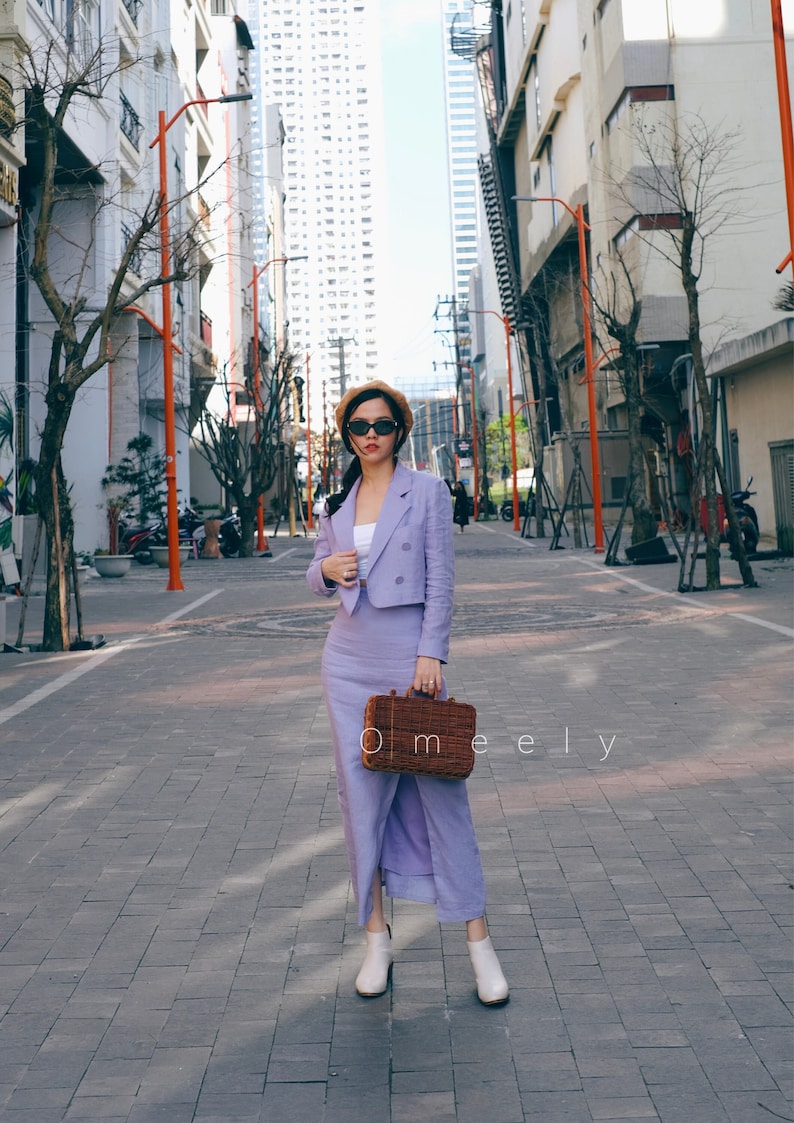 Pu&ograve; includere: Un completo due pezzi color lavanda composto da un blazer corto a doppio petto e una gonna lunga abbinata, indossato con un top bianco, un basco beige, occhiali da sole neri, stivaletti bianchi e una borsa in vimini marrone con la scritta Omeely.