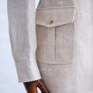 May include: A beige linen jacket featuring a long sleeve with a buttoned cuff and a large patch pocket on the side with a flap and a single button closure. The word Omeely is printed in white text on the lower portion of the jacket fabric.