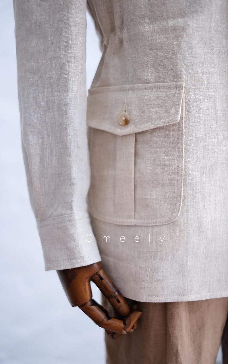 May include: A beige linen jacket featuring a long sleeve with a buttoned cuff and a large patch pocket on the side with a flap and a single button closure. The word Omeely is printed in white text on the lower portion of the jacket fabric.