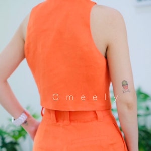 K&ouml;nnte beinhalten: Ein leuchtend orangefarbenes Leinen-Crop-Top und eine passende, hoch taillierte Hose. Das Top hat einen Kragen und ein &auml;rmelloses Design. Die Hose hat eine weite Beinweite und G&uuml;rtelschlaufen. Das Model tr&auml;gt das Outfit und hat ein kleines Kaktus-Tattoo auf dem Arm.