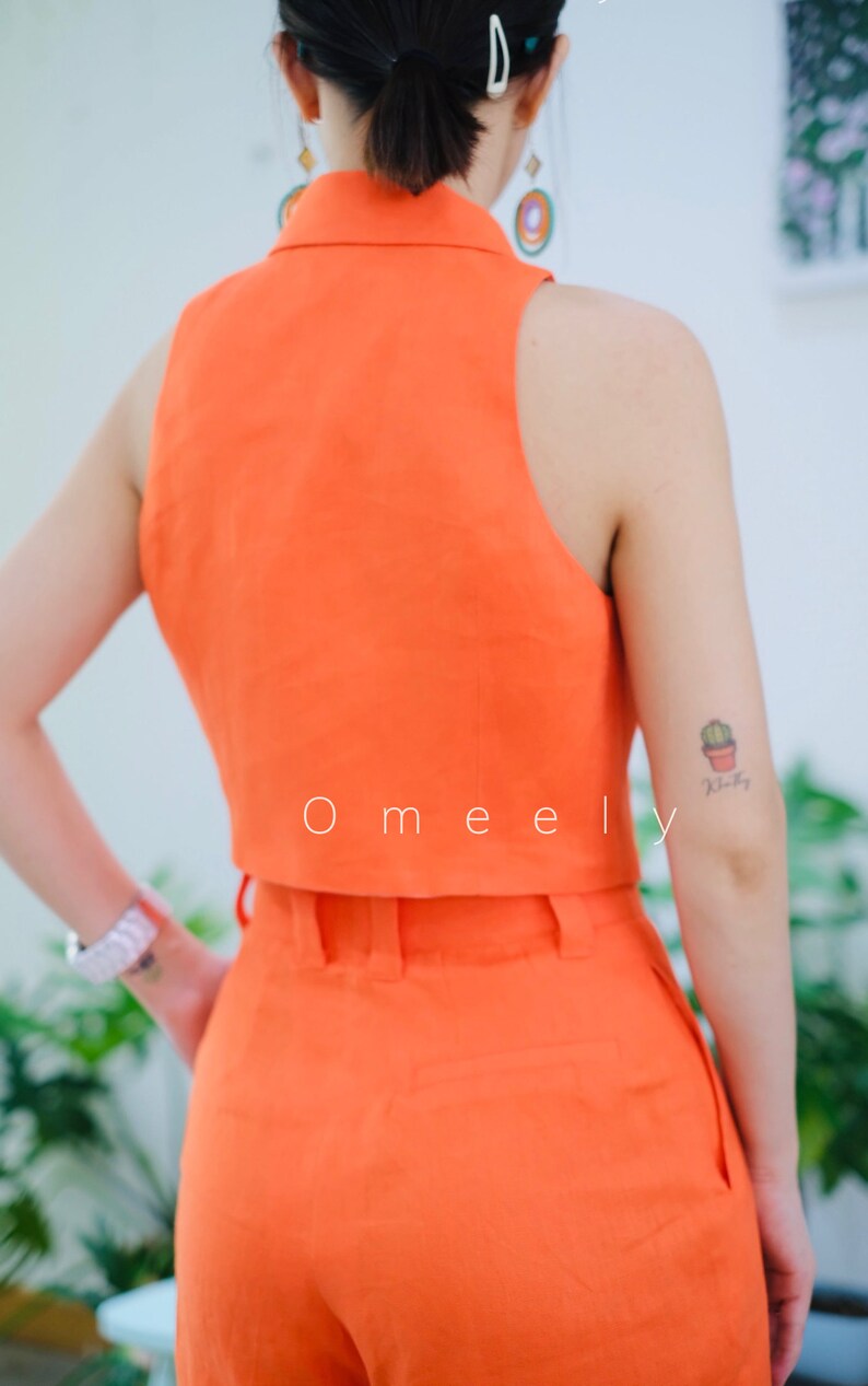 K&ouml;nnte beinhalten: Ein leuchtend orangefarbenes Leinen-Crop-Top und eine passende, hoch taillierte Hose. Das Top hat einen Kragen und ein &auml;rmelloses Design. Die Hose hat eine weite Beinweite und G&uuml;rtelschlaufen. Das Model tr&auml;gt das Outfit und hat ein kleines Kaktus-Tattoo auf dem Arm.