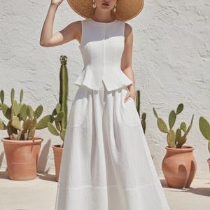 May include: A woman wearing a white sleeveless top and a matching maxi skirt. She is also wearing a wide-brimmed straw hat and white sandals. The background features potted cacti and a textured white wall.