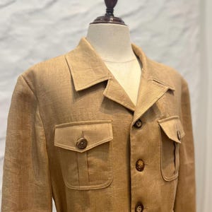 May include: A tan linen jacket featuring a collared neckline, long sleeves, and a front button closure. The garment includes two chest pockets with buttoned flaps and a cinched waist detail. The word Omeely is printed in white on the lower right.
