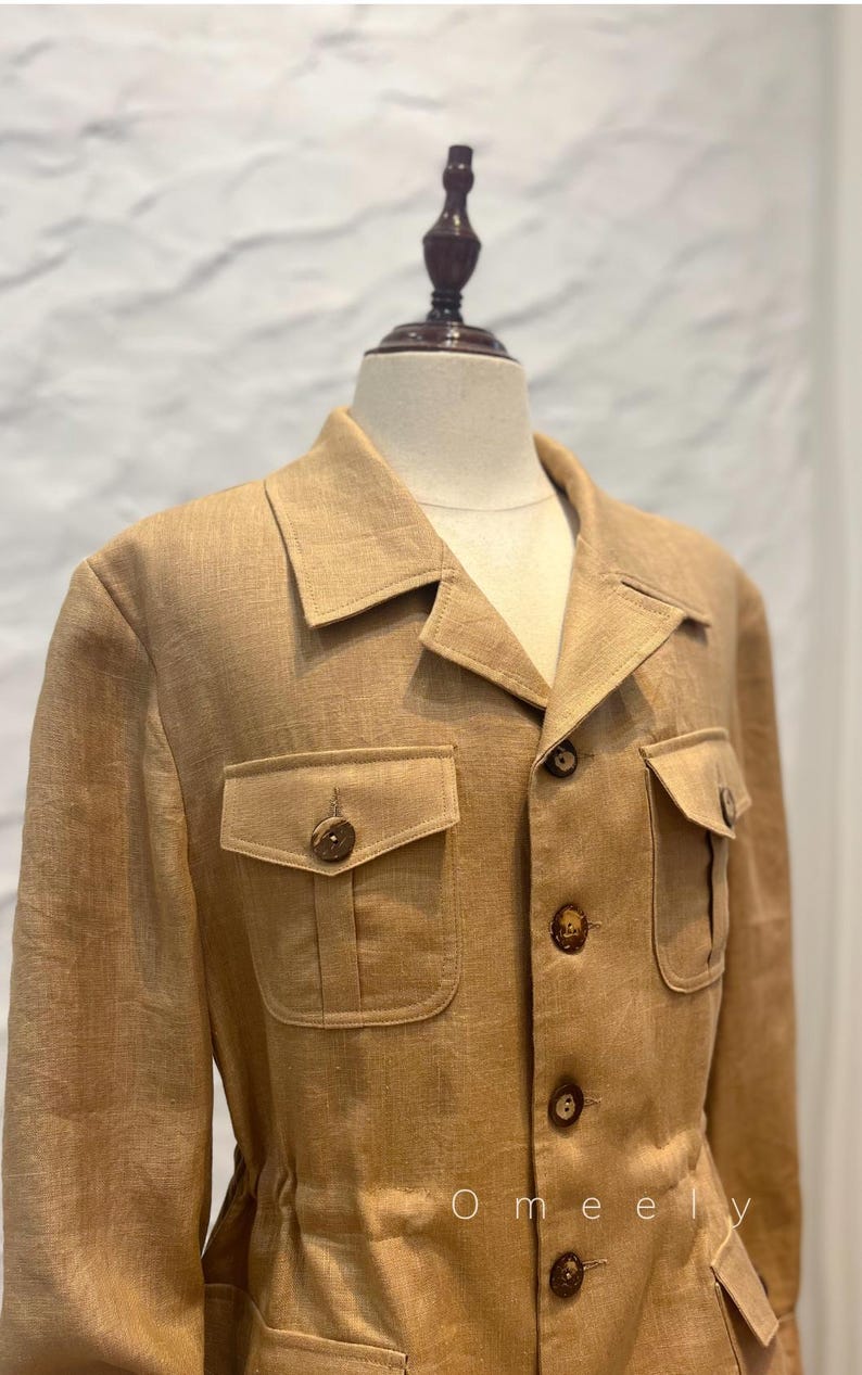 May include: A tan linen jacket featuring a collared neckline, long sleeves, and a front button closure. The garment includes two chest pockets with buttoned flaps and a cinched waist detail. The word Omeely is printed in white on the lower right.