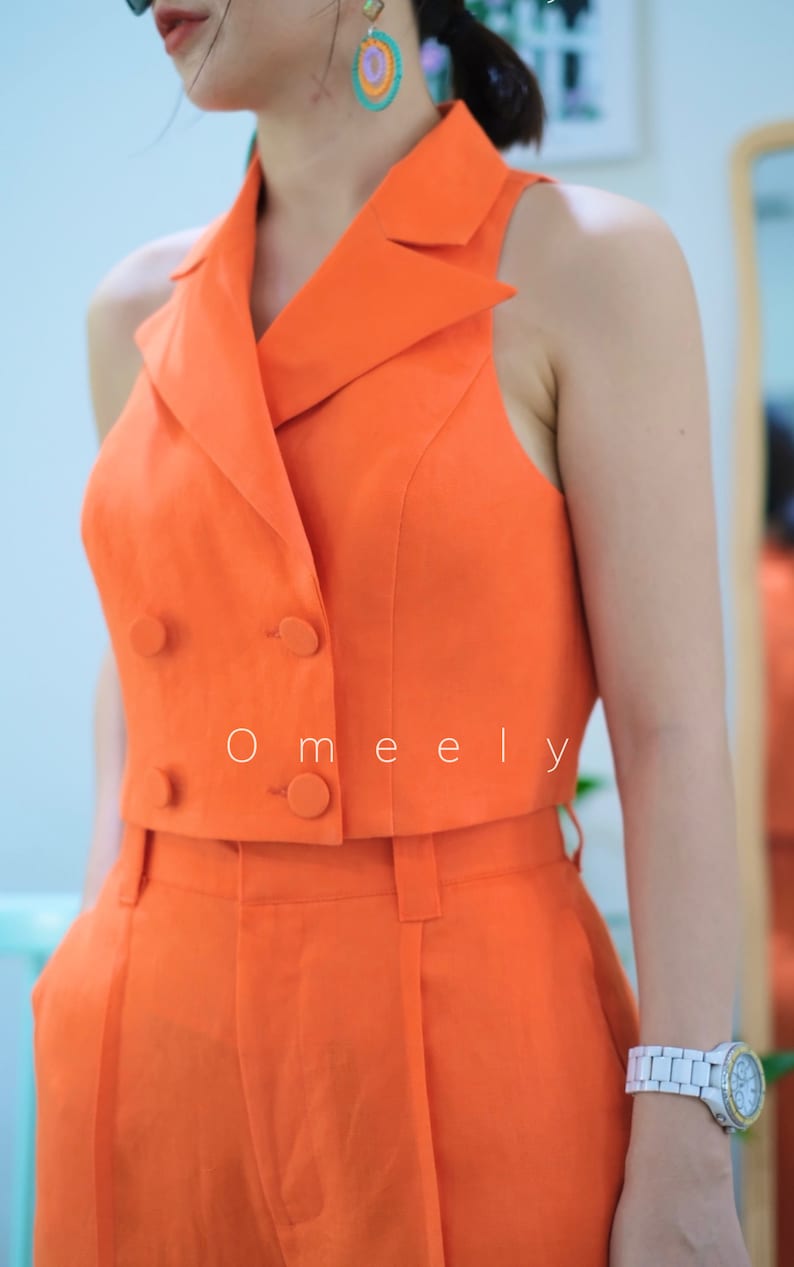 May include: A sleeveless orange cropped blazer with a double breasted closure and two buttons. The blazer is paired with matching high waisted orange pants. The text 'O meely' is visible on the front of the blazer.