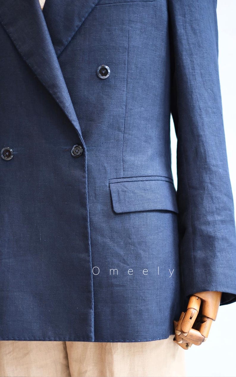 Op de afbeelding: Een marineblauwe double-breasted blazer met een ingekeepte revers, lange mouwen en een enkele klepzak aan de voorzijde. Het kledingstuk heeft donkere knopen en de tekst Omeely in het wit gedrukt op het onderste gedeelte van de stof.