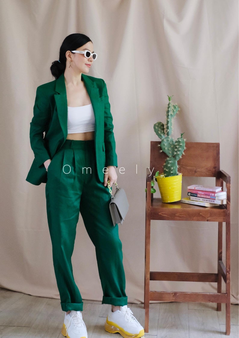 May include: A woman wearing a green suit with a white top and white trainers. She is holding a grey clutch bag with a chain strap. The woman is standing in front of a wall with a cactus plant in a yellow pot on a wooden stool.